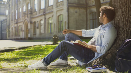 Multinational guy sitting under tree with book, looking sideways, leisure time