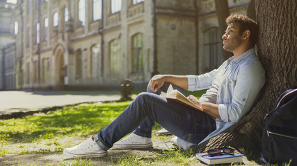 Young male sitting under tree with book looking around, having pleasant thoughts