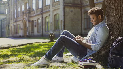 Multiracial young man sitting under tree, scrolling mobile phone screen, smiling