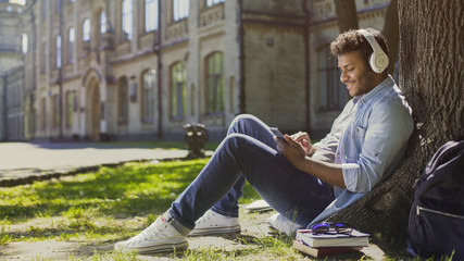 Obraz premium Young male in headphones sitting on grass, leaning against tree, choosing song
