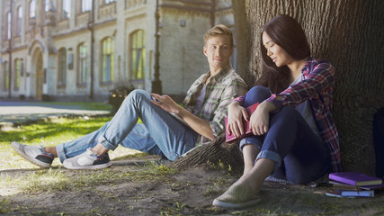 Two young people sitting under tree, girl looking down and guy looking at her