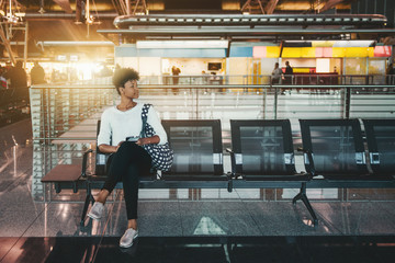 Lonely Brazilian girl is sitting inside of modern airport terminal on metal seats and waiting her flight; cute young African American female tourist is waiting for her train indoors of railway station