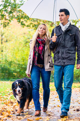 Naklejka na ściany i meble Woman and man having walk with dog in autumn rain in park