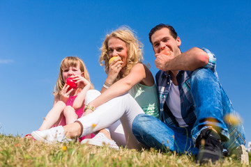 Mommy, daddy and little girl sitting on meadow eating fruit