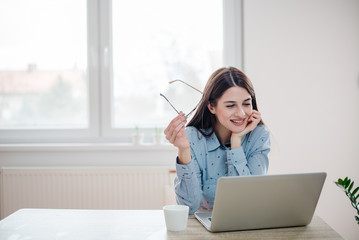 Young woman holding her eyeglasses while sitting in front of laptop computer.