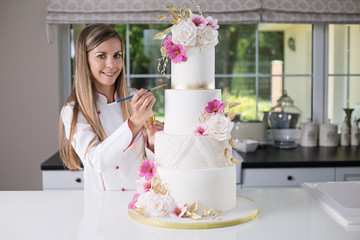 Beautiful smiling confident confectioner in chefs uniform standing in the modern kitchen, holding a brush and gold paint, decorating a four tier white, pink and gold wedding cake