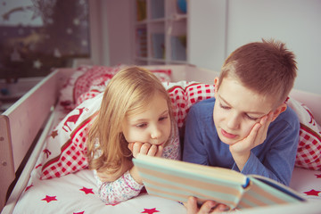 Fototapeta premium Two happy sibling children reading book in bunk bed under blanket