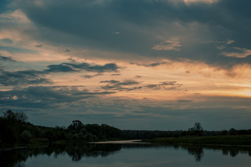 clouds and forest by the river on a meadow at sunset. Rural landscape.