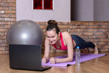 A young woman performs a plank exercise on a purple mat on parquet floor and watching instructional...