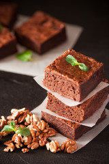 Chocolate brownie square pieces in stack on white plate with walnuts, decorated with mint leaves and cocoa on black background. Delicious dessert. Dark mood. Close up photography. Selective focus