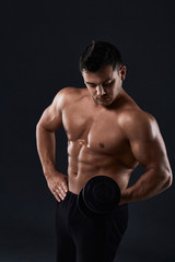 Naklejka na ściany i meble Muscular bodybuilder doing exercises with dumbbell over black background.Strong athletic man shows body,abdominal muscles,biceps and triceps.Work out,gaining weight,pumping up muscles with dumbbells.