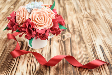 Hand made bouquet from foamiran beige roses, red alstroemeria and white chrysanthemums with red satin ribbon on wooden background with copy space.