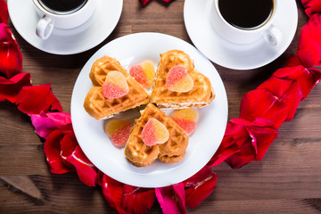 On the wooden table, inside the heart of the rose petals are two cups of coffee and a plate with biscuits. The middle plan