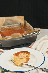 Freshly baked, homemade, warm apple buckle coffee cake, round, white plate, blue rim, artisan fork, printed towel, rest of cake in metal baking pan cooling on metal trivet