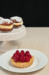 Freshly prepared, mini, raspberry cream tart on a round, white plate with two white cream filled donuts on a pedestal white, round display dish