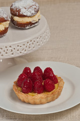 Freshly prepared, mini, raspberry cream tart on a round, white plate with two white cream filled donuts on a pedestal white, round display dish
