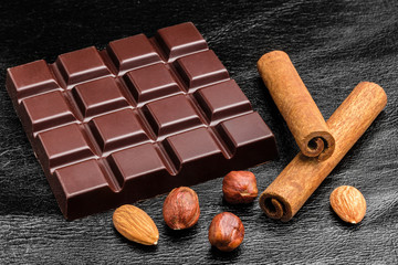 Dark, bitter chocolate bar, almond nuts, hazelnut and two cinnamon sticks on black leather texture...