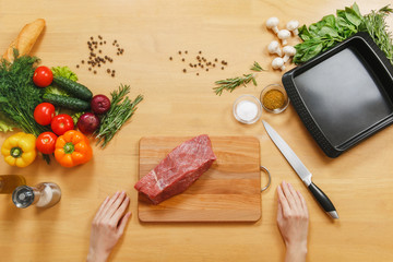 Raw meat. Fresh beef tenderloin on cutting board on wooden table with different vegetables, spices, mushrooms, baking tray, knife. Top view flat lay Copy space for advertisement. Woman cuts with knife