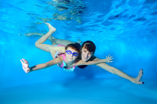 Mom and daughter swim together underwater in the pool, spread wide his arms to the side, looking at the camera and smiling. Portrait. Horizontal orientation. The view from under the water