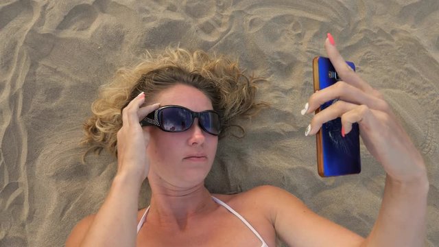 Woman with cell phone on the beach. She lies on bare sand. The girl rewrites communicates on the phone, does selfie and looks at the finished pictures.