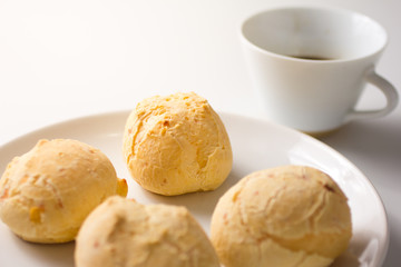 Pao de Queijo is a cheese bread ball from Brazil. Also known as Chipa, Pandebono and Pan de Yuca. Snacks on white plate and cup of coffee.