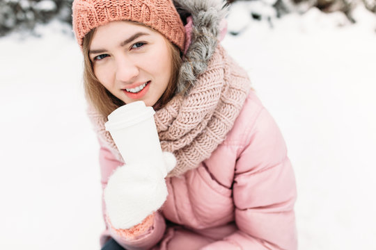 Portrait of a beautiful young girl in white knitted mittens, outdoors, holding a paper Cup with a hot beverage, bright winter day.the woman smiles and is happy, close-up, Trees in first snow in the.