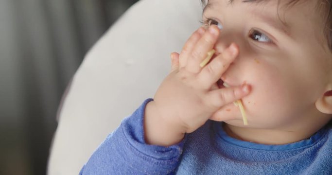Baby Eats Spaghetti and Looks Up . a close up view of a baby in a highchair eating pasta with his hands and licking fingers. Slow motion