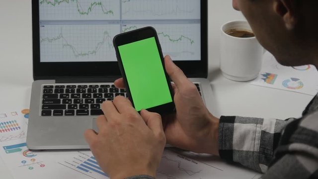Businessman using smartphone with green screen at white office table background. Top view. Male hands scrolling pages, zooming, tapping on touch screen. Office desk background