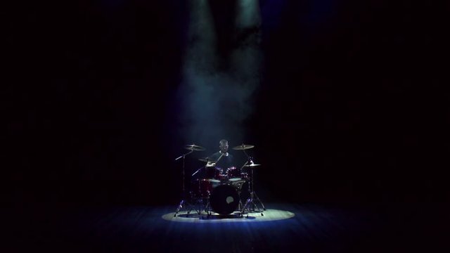 A young bearded man plays drums in the dark on stage during a concert, slow motion. The drummer is enjoying his performance during the concert. Man playing drums on black background with smoke.