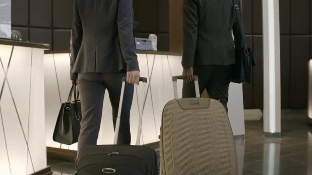 Rear view of businessman and businesswoman pulling suitcases and walking to check–in desk. Friendly female receptionist greeting guests and talking with them
