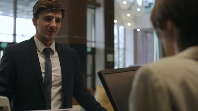 Young businessman standing at check-in desk in hotel lobby and talking with female receptionist