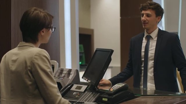 Young businessman speaking with female receptionist at check-in desk in hotel and paying for room with smartphone