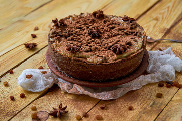Homemade cheesecake with chocolate powder on the brown plate decorated with white cloth, silver spoon and raisins. Powdered chocolate tart with anise stars on the wooden background. Side view