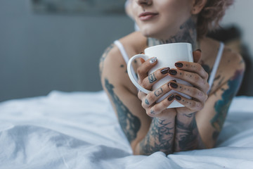 cropped view of tattooed girl drinking coffee in bed at morning