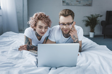 tattooed smiling couple using laptop together in bed