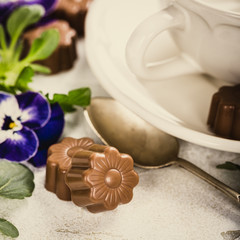 Chocolate candies in flower shape with violets and cup of coffee. Holiday food concept with copy space. Retro style toned postcard.