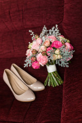 Wedding bouquet with purple and pink roses lying on a red armchair. Beige bridal shoes out of focus.