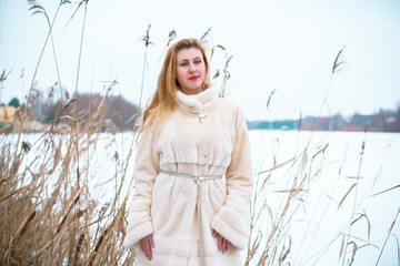 Fototapeta na wymiar Beautiful woman in a white mink coat stands against the background of a snowy winter landscape, white field. Young woman of European or American appearance is refined and elegant in white furs 