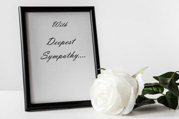 White condolence card with text. Black frame and white artificial rose on the light background. Sorrow concept.
