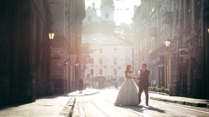 Full-length wedding shot of the stylishly dressed newlywed couple talking and holding hands during their walk along the road of the city centre.