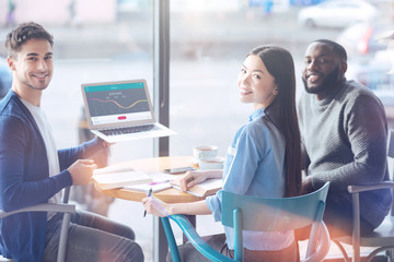 Look here. Handsome smiling man sitting in semi position and keeping laptop in left hand while posing on camera