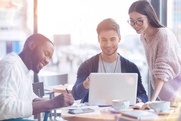 Work in one group. Handsome bearded male person looking at screen of computer and keeping smile on his face while feeling happiness