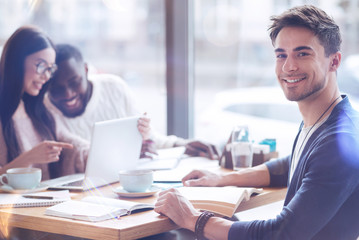 Share your positivity. Handsome man smiling while going to read interesting book and sitting opposite his best friends