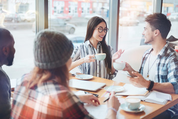 Tell it us. Pretty longhaired international girl sitting opposite her colleague, looking at him while holding cup of tea in right hand