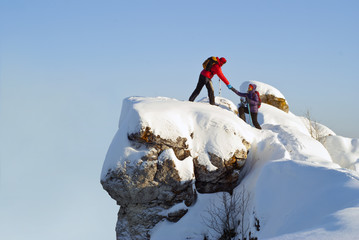 two hikers on top of the mountain in winter; a man helps a woman to climb a sheer stone