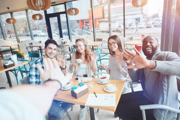 Hey you. Positive delighted young people sitting together in cafe and waving hands while looking straight at camera