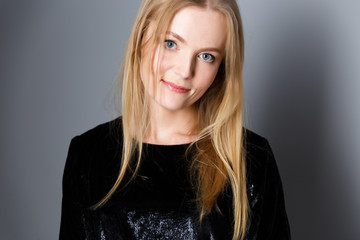 portrait of a young beautiful blond woman with evening make-up in a black shiny dress near a gray wall