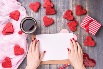 Valentine's day love letter on wooden background. Brown envelop, pink note and gift box on table. Red velvet heart shape cookies, candy and coffee. Female hands with red nail polish, manicure