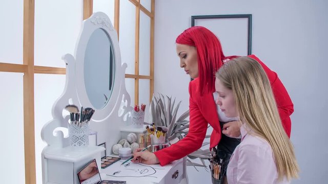 A make up artist shows the client how she will draw her eyebrows. She shows her on an example in the picture.