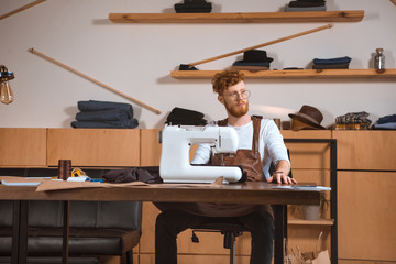 Fototapeta premium handsome young fashion designer in eyeglasses looking away while working with sewing machine in workshop